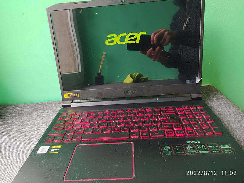 Acer Nitro 5 ноутбук з увімкненим екраном і відображенням на ньому логотипу Acer, червоною підсвіткою клавіатури та технічними стікерами на панелі біля трекпаду. На фоні видно зелений стіл та людину, яка тримає телефон для фотографування екрану.
