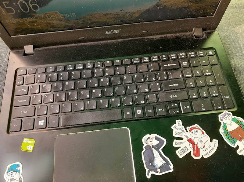 На цьому зображенні показано ноутбук Acer з російською розкладкою клавіатури, прикрашений наклейками. Екран увімкнений, на ньому відображається заставка з часом 5:06 і датою "Вівторок, 7 березня". Ноутбук має класичний дизайн із чорним корпусом і стандартною клавіатурою для ноутбуків цього бренду.