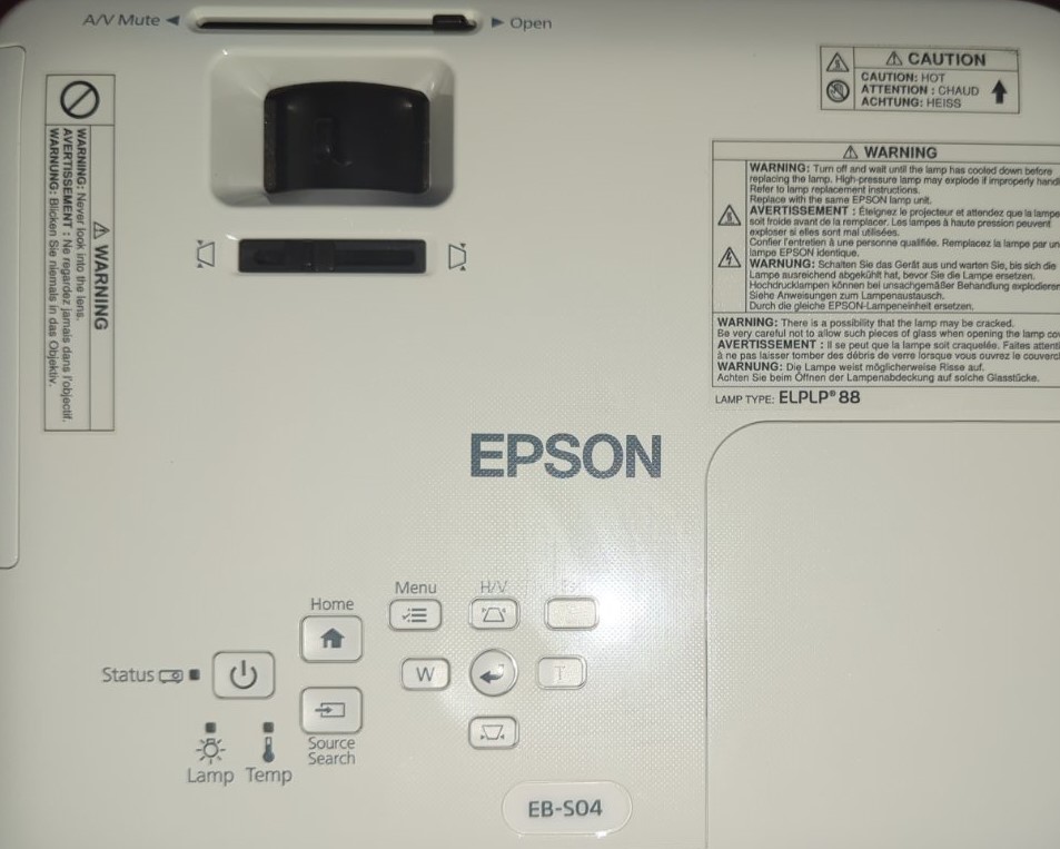 epson-projector-top-view-controls.jpg Вид зверху проектора Epson, модель EB-S04, з кнопками управління, наклейками з попередженнями та налаштуваннями фокуса.