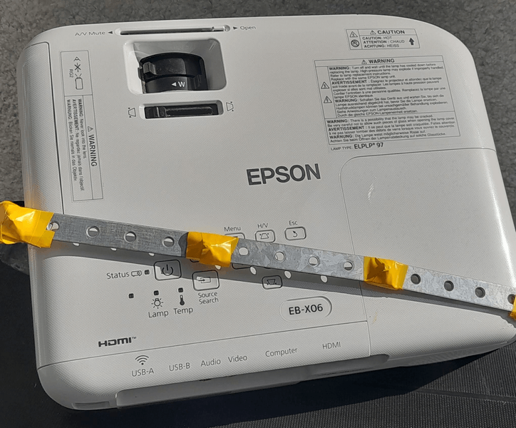 Проектор Epson EB-X06 з індикаторами та попередженнями. Проектор Epson EB-X06 з індикаторами стану та кнопками керування, включаючи HDMI, USB, AV Mute, і джерело пошуку. Видима наклейка про попередження щодо електричної безпеки.