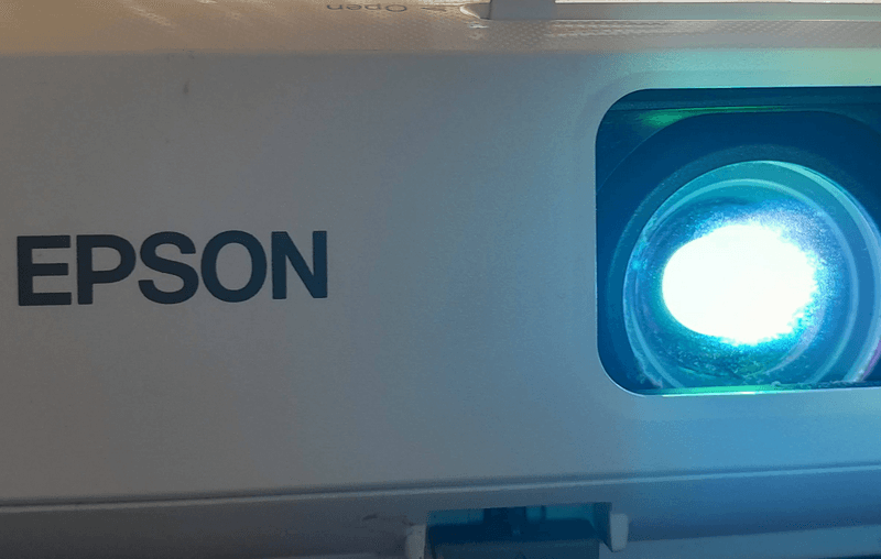 Проектор Epson з увімкненим об'єктивом Проектор Epson під час роботи, на зображенні чітко видно його об'єктив, через який проходить яскраве світло. Бренд Epson добре помітний на корпусі проектора.