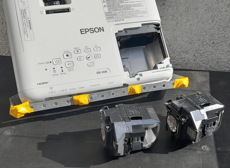Проектор Epson EB-X06 з вийнятими лампами для ремонту Задня частина проектора Epson, модель EB-X06, з двома вийнятими лампами, що лежать поруч. Проектор закріплений на металевій пластині, обмотаній жовтою ізоляційною стрічкою, ймовірно, для ремонту або модифікації.